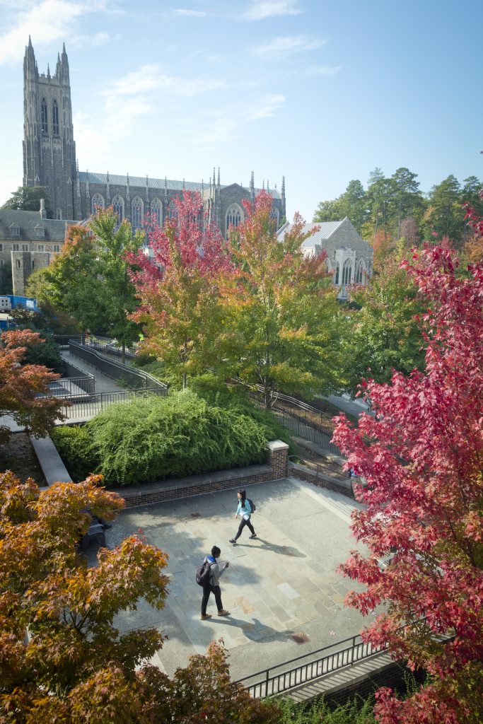 Students walk across the Bostock Library plaza as the Chapel is visible in the distance above the colorful fall trees.